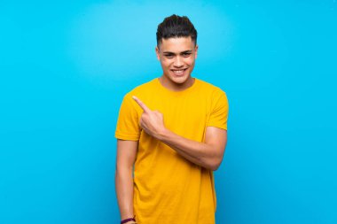 Izole mavi arka plan üzerinde sarı gömlek li genç adam bir ürün sunmak için yan işaret