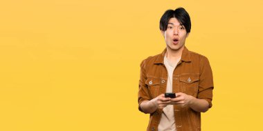 Kahverengi ceketli Asyalı adam şaşırttı ve izole sarı arka plan üzerinde bir mesaj gönderme