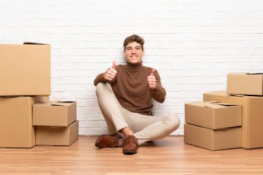 Yakışıklı genç adam kutular arasında yeni bir evde hareket jest bir başparmak veren