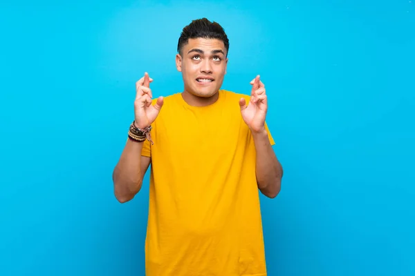 Young man with yellow shirt over isolated blue background with fingers crossing and wishing the best