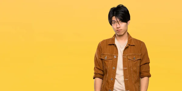 Asian man with brown jacket with sad and depressed expression over isolated yellow background