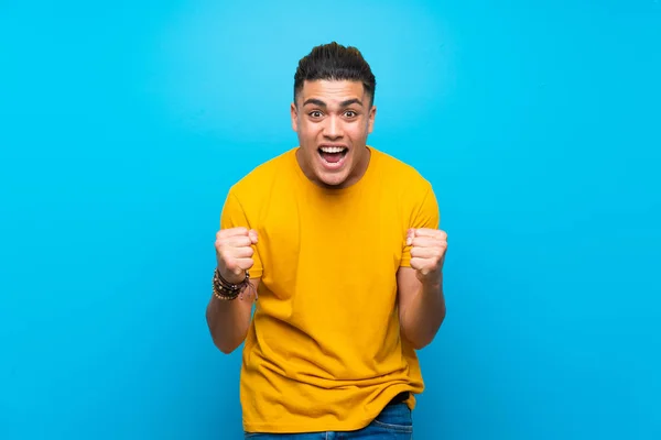 Young man with yellow shirt over isolated blue background celebrating a victory in winner position