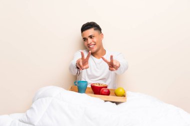 Yatakta kahvaltı edip gülümseyen ve zafer işareti gösteren genç adam