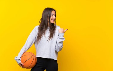 Genç kadın bir ürün sunmak için yan işaret izole sarı arka plan üzerinde basketbol oynuyor