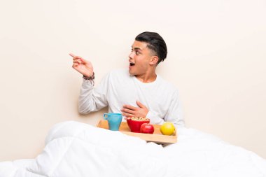Yatakta kahvaltı eden genç adam parmağını yana doğru gösteriyor