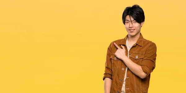 孤立した黄色の背景の上に製品を提示するために側面を指す茶色のジャケットを持つアジアの男性 — ストック写真