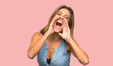 Jean elbiseli sarışın kadın bağırıyor ve izole pembe arka plan üzerinde bir şey ilan