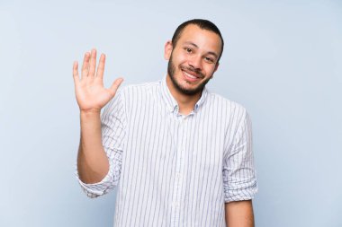 Mutlu ifade ile el ile selamlama izole mavi duvar üzerinde Kolombiyalı adam