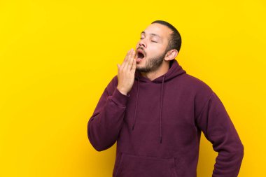 Sarı duvar esneme üzerinde sweatshirt ile Kolombiyalı adam esneme ve el ile geniş açık ağız kapsayan