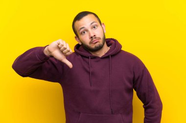 Olumsuz ifade ile başparmak aşağı gösteren sarı duvar üzerinde sweatshirt ile Kolombiyalı adam