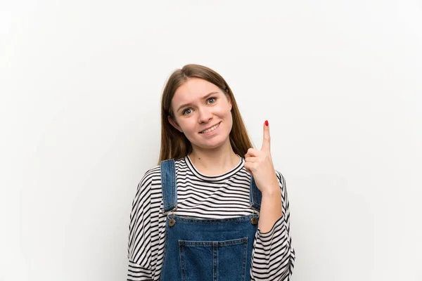 Young woman in dungarees over white wall pointing with the index finger a great idea