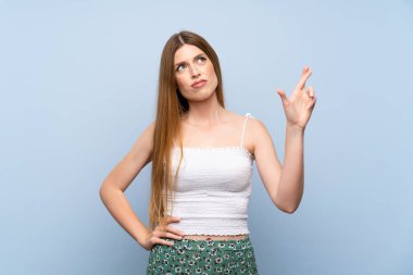 İzole edilmiş mavi arka planı olan genç bir kadın parmaklarını çapraz yapıp en iyisini diliyor.