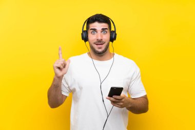 Genç yakışıklı adam izole sarı duvar üzerinde bir cep ile müzik dinlerken harika bir fikir işaret