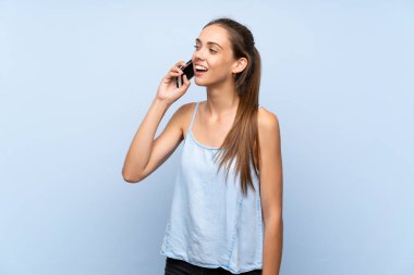 İzole edilmiş mavi arka planı olan genç bir kadın cep telefonuyla sohbet ediyor.