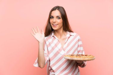 Genç kadın mutlu ifade ile el ile selamlama izole pembe duvar üzerinde bir pizza tutan