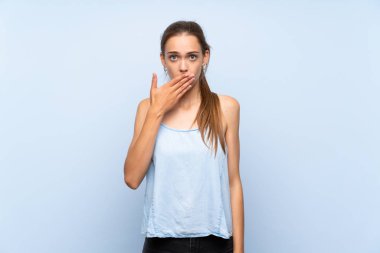 İzole edilmiş mavi arka planı olan genç bir kadın elleriyle ağzını kapatıyor.