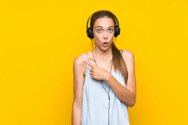 Mujer Joven Escuchando Música Sobre Aislada Pared Amarilla Sorprendida Señalando —  Fotos de Stock