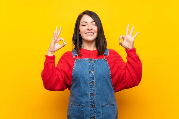 Young Mexican woman with overalls over yellow wall in zen pose
