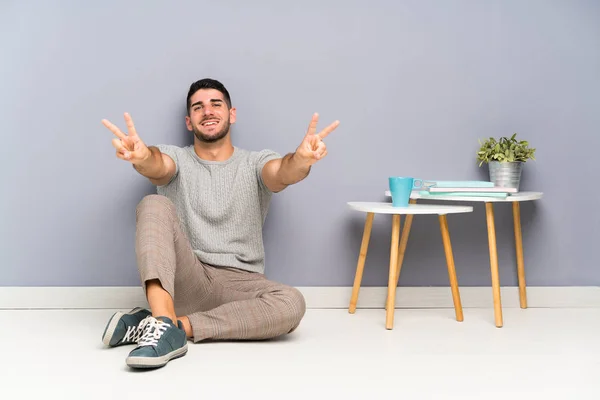 Jovem Homem Bonito Sentado Chão Sorrindo Mostrando Sinal Vitória — Fotografia de Stock