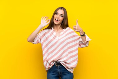 İzole edilmiş sarı arka plan üzerinde genç bir kadın tamam işareti ve baş parmak işareti gösteriyor.