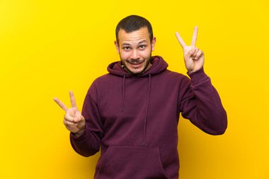 Sarı duvar üzerinde sweatshirt ile Kolombiyalı adam iki eliyle zafer işareti gösteren