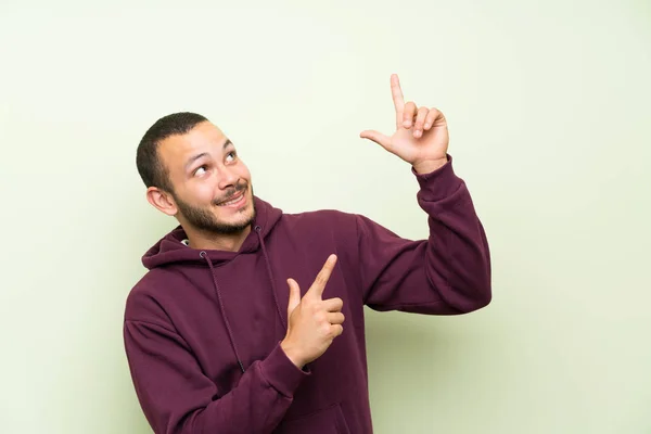 Hombre Colombiano Con Sudadera Sobre Pared Verde Señalando Con Dedo —  Fotos de Stock