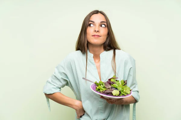 Young woman with salad over isolated green wall making doubts gesture while lifting the shoulders