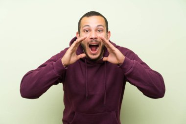 Yeşil duvar üzerinde Sweatshirt ile Kolombiyalı adam ağız geniş açık bağırarak