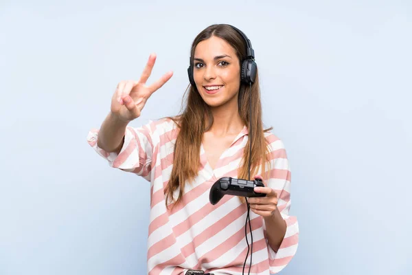 年轻女子玩视频游戏控制器在孤立的蓝色墙壁微笑 并显示胜利标志 — 图库照片
