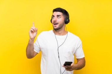 Genç yakışıklı adam izole sarı duvar üzerinde bir cep ile müzik dinlerken bir parmak kaldırırken çözüm gerçekleştirmek niyetinde