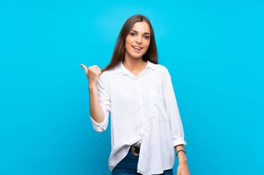 İzole edilmiş mavi arka planda genç bir kadın bir ürünü sunmak için kenarı işaret ediyor.