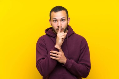 Sarı duvar üzerinde sweatshirt ile Kolombiyalı adam ağzına parmak koyarak sessizlik jest bir işareti gösteren