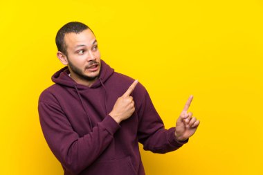 Sarı duvar üzerinde sweatshirt ile Kolombiyalı adam korkmuş ve yan işaret