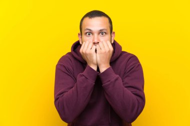 Sarı duvar üzerinde sweatshirt ile Kolombiyalı adam sinirli ve ağız ellerini koyarak korkmuş