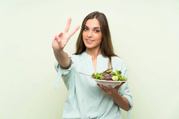 Young woman with salad over isolated green wall smiling and showing victory sign