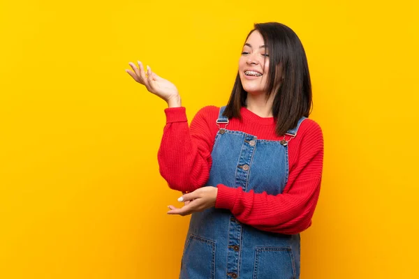 Young Mexican woman with overalls over yellow wall extending hands to the side for inviting to come