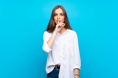 İzole edilmiş mavi arka planda genç bir kadın sessizlik işareti gösteriyor. Parmağını ağzına sok.