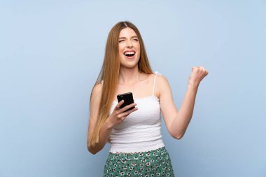 İzole edilmiş mavi arka planı olan genç bir kadın ve telefon zafer pozisyonunda.