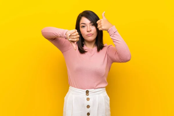 Young Mexican woman over isolated yellow background making good-bad sign. Undecided between yes or not