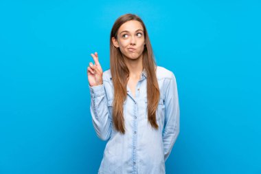 İzole edilmiş mavi arka planı olan genç bir kadın parmaklarını çapraz yapıp en iyisini diliyor.