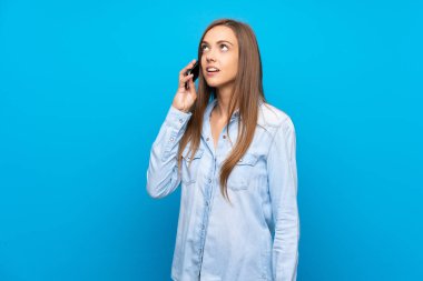 İzole edilmiş mavi arka planı olan genç bir kadın cep telefonuyla sohbet ediyor.