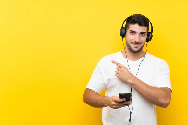 孤立した黄色の壁の上にモバイルで音楽を聴いている若いハンサムな男は 製品を提示するために側面を指し示す — ストック写真