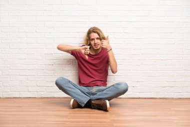 Sarışın adam yerde oturmuş iyi-kötü işareti yapıyor. Evet ya da değil arasında kararsız