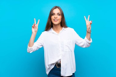 İzole edilmiş mavi arka planda genç bir kadın zafer işaretini iki eliyle gösteriyor.