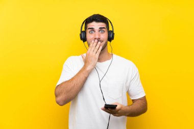 Genç yakışıklı adam sürpriz yüz ifadesi ile izole sarı duvar üzerinde bir cep ile müzik dinliyor