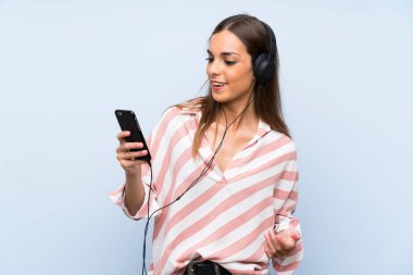 İzole edilmiş mavi duvarda cep telefonu ile müzik dinleyen genç bir kadın.