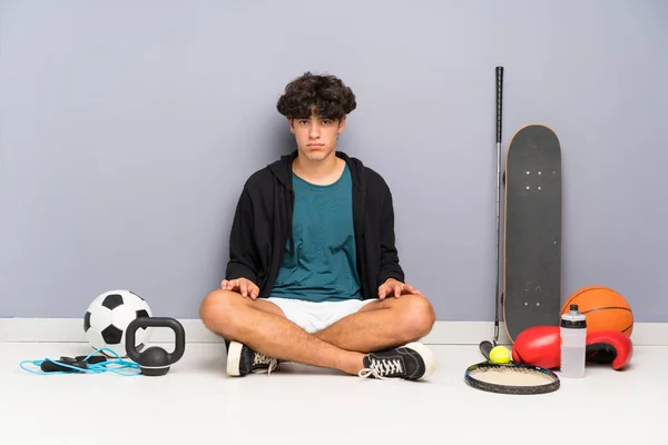 Young sport man sitting on the floor around many sport elements sad