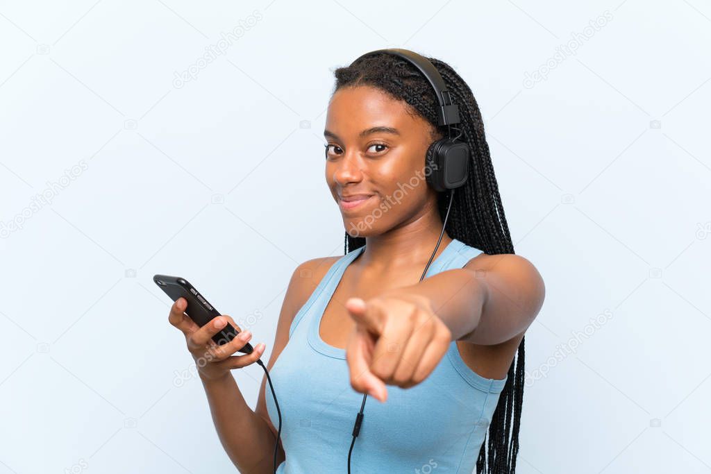 African American teenager girl with long braided hair listening music with a mobile points finger at you with a confident expression