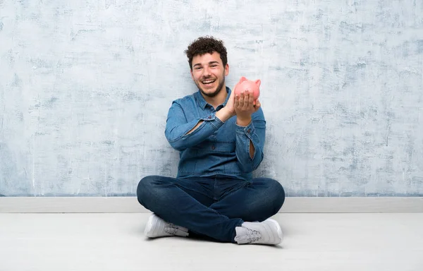 Yerde oturan genç adam elinde büyük bir kumbarayla