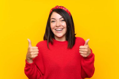 Sarı duvar üzerinde kırmızı kazak ile genç Meksikalı kadın jest kadar bir başparmak veren